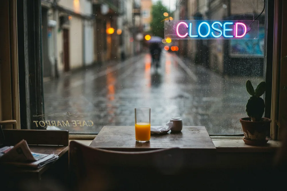 The same cafe window after three Ideogram 4.5 edits: the sign reads CLOSED in the original pink and blue, a glass of orange juice sits on the table and a small cactus replaces the fern