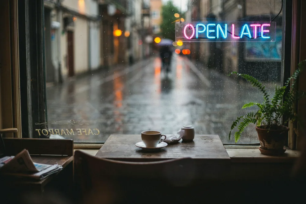 A rainy cafe window with a pink and blue neon sign reading OPEN LATE, a coffee cup on a wooden table and a fern on the windowsill, generated by Ideogram 4.5